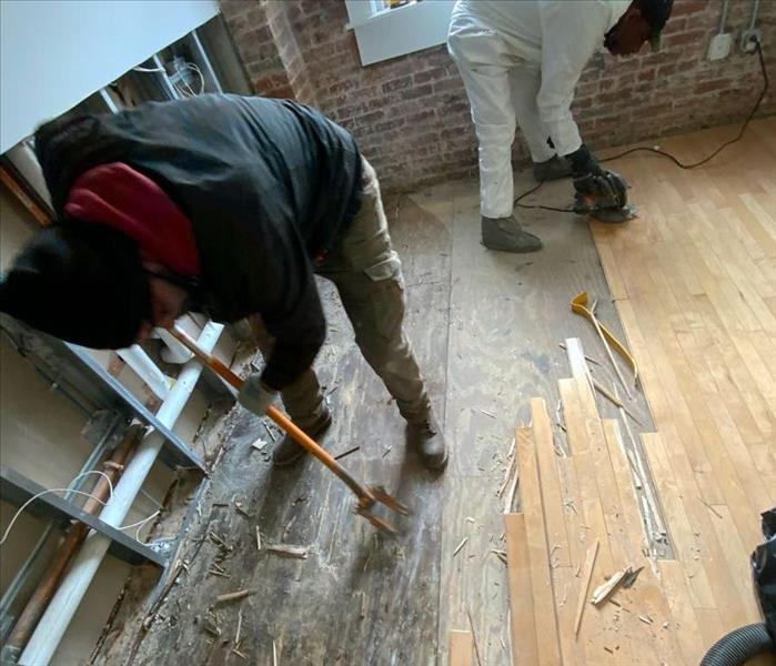 Two SERVPRO technicians sand and pull up floorboards and expose the wet subfloor underneath near a demolished office wall