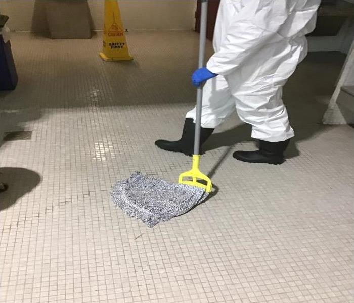 technician in trec suit and lack rubber boots mopping wet bathroom floor in commercial building